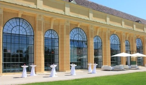 Come sfondo si vede l’Orangerie di Vienna con le finestre arcuate, nel piazzale antistante nel giardino con tavolini e lounge, ideali per un aperitivo prima del concerto dell’orchestra del castello di Schönbrunn.