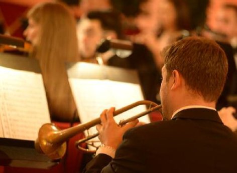 シェーンブルン宮殿劇場管弦楽団の管楽器楽者がトランペットを演奏しています。後方に楽譜やオーケストラの他演奏家たちがぼんやりと見えます。