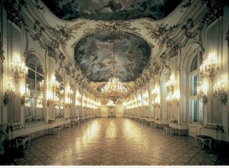 Una sala suntuosa en el Palacio de Schönbrunn; a la izquierda y derecha en las paredes alumbran varios candelabros, en el techo pueden verse pinturas fastuosas